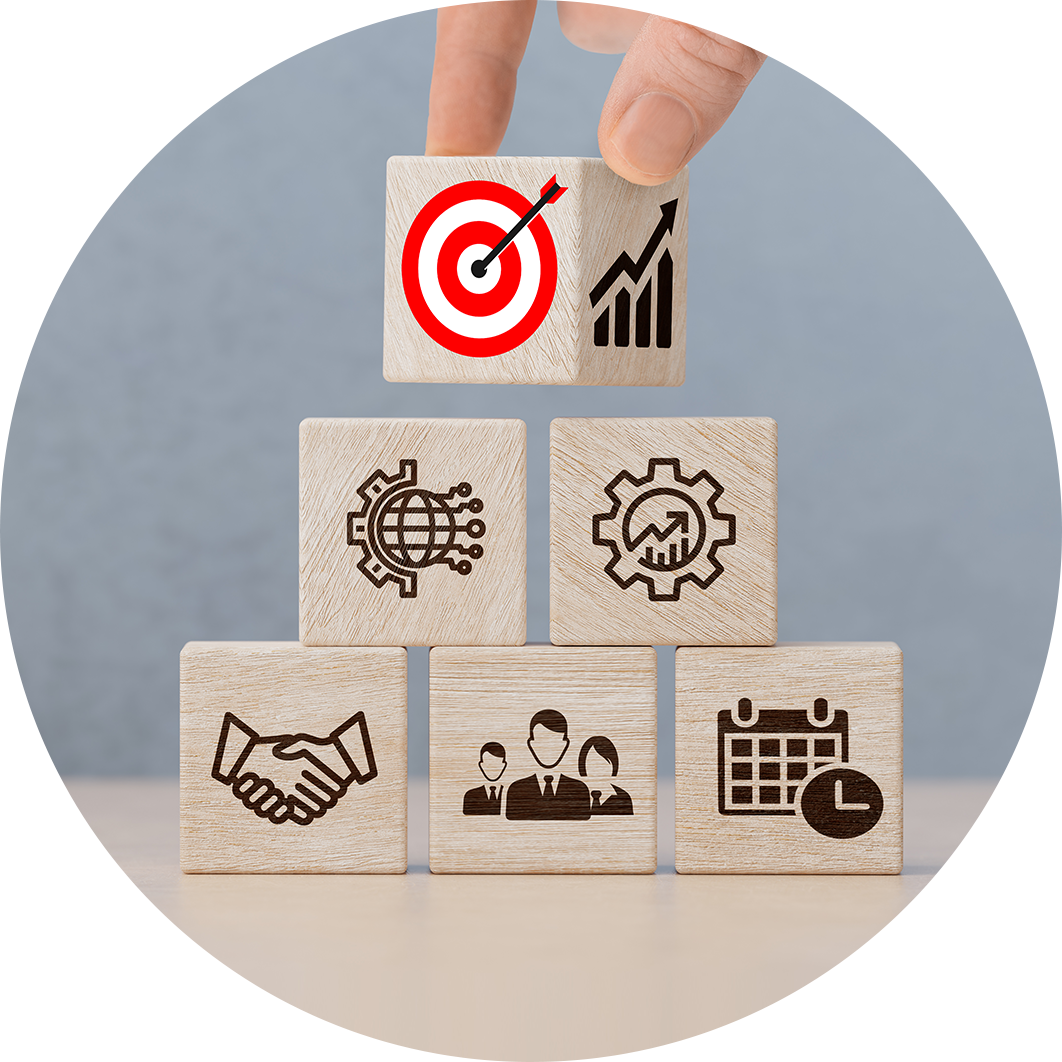 Eine Hand platziert einen Block mit einem Zielsymbol auf einer Pyramide aus gesch&auml;ftsbezogenen Symbolen auf Holzw&uuml;rfeln. Bild von der EMIL-Akademie f&uuml;r Emotionale Intelligenz & Leadership vor Ort in K&ouml;ln & Online. Emotionale Intelligenz & F&uuml;hrung auch durch Mimikresonanz, Mesource Design Thinking, Integrale Organisationsentwicklung und Facilitation f&uuml;r Fach- & F&uuml;hrungskr&auml;fte.